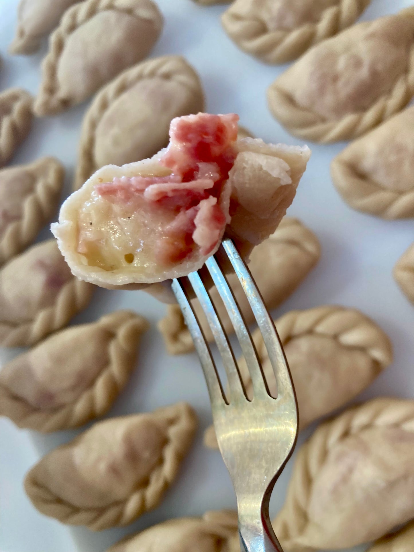 A close-up image of a fork holding a piece of bacon and cheddar cheese filled potato pierogi, with more pierogi in the background.